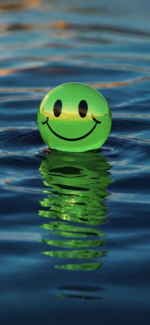 Green smiley face ball floating on calm water, reflecting vibrant color. Serene, joyful vibe. | 4K Wallpaper for Mobile