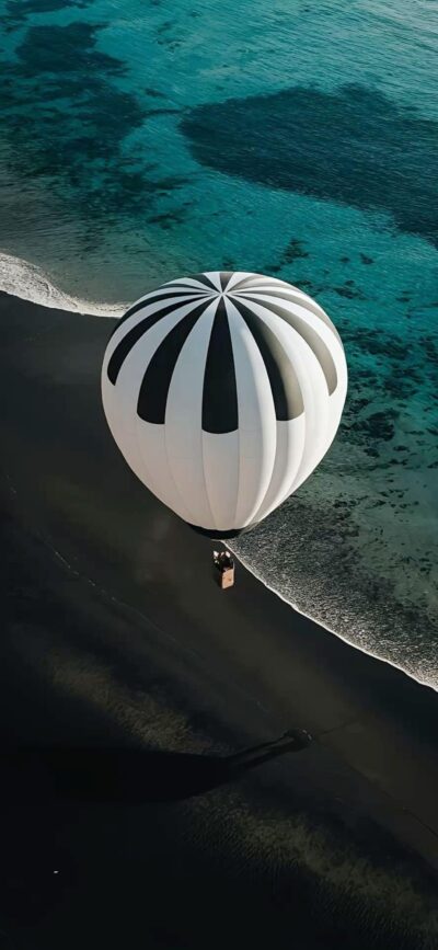 Hot air balloon with black and white stripes over a coastline, blue ocean, and dark sandy beach | 4K Wallpaper for Mobile