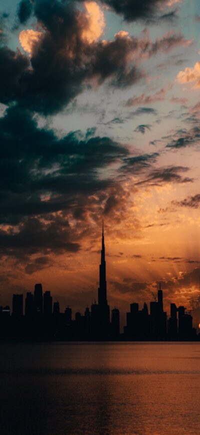 Cityscape skyline silhouetted at sunset with dramatic clouds, orange and yellow hues | 4K Wallpaper for Mobile